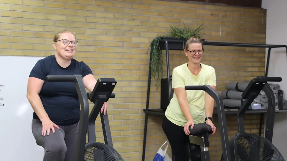 To kvinder træner på motionscykler i Box One's træningshal med gule mursten i baggrunden